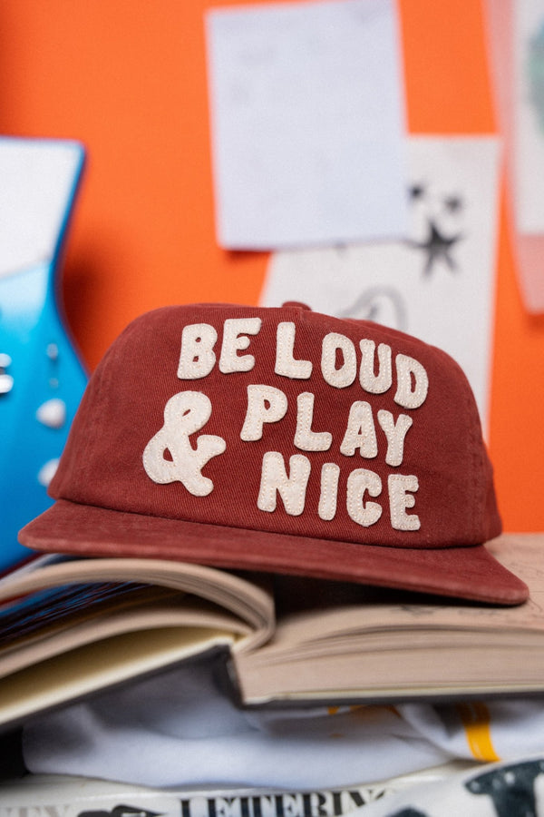 Red cap with 'Be Loud & Play Nice' text on a stack of books with an orange background