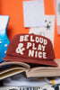 Red cap with 'Be Loud & Play Nice' text on a stack of books with an orange background