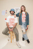 Two people wearing matching t-shirts with text and graphics that read "live band every night musicians drink free" sitting on a stool against a plain background.