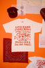 White t-shirt with red text and graphics that read "live band every night musicians drink free"on an orange background, accompanied by a bandana and another t-shirt.