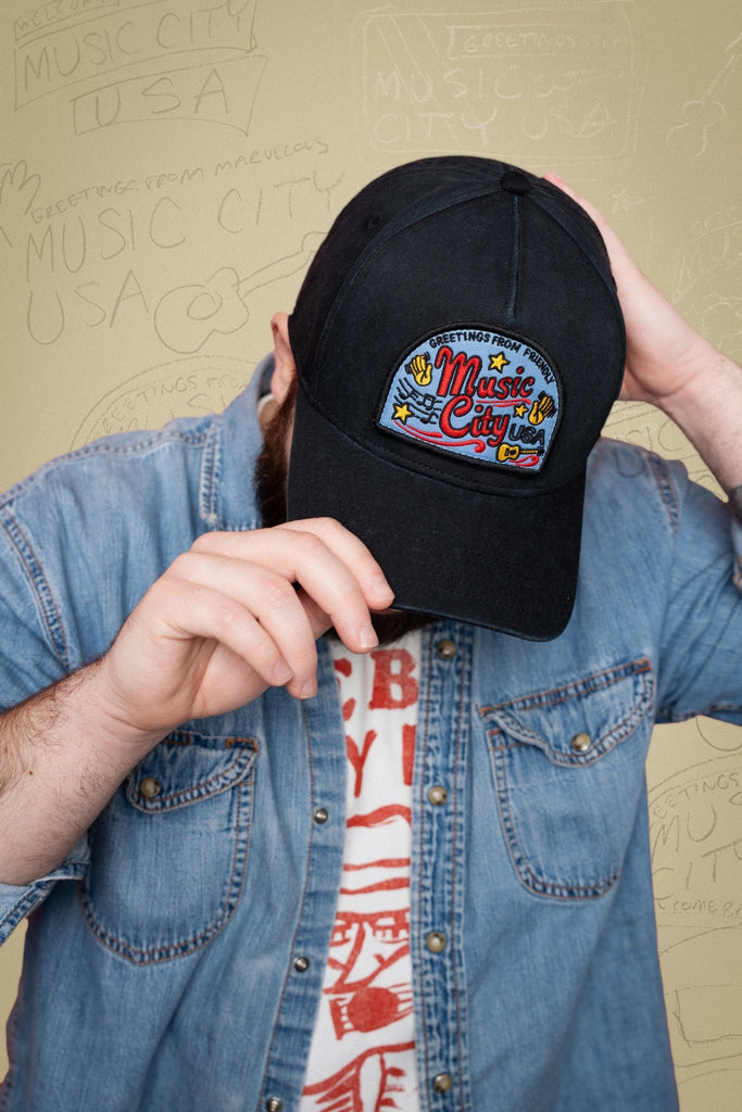 Person wearing a black cap with a colorful 'Music City USA' design, holding it up to their face.