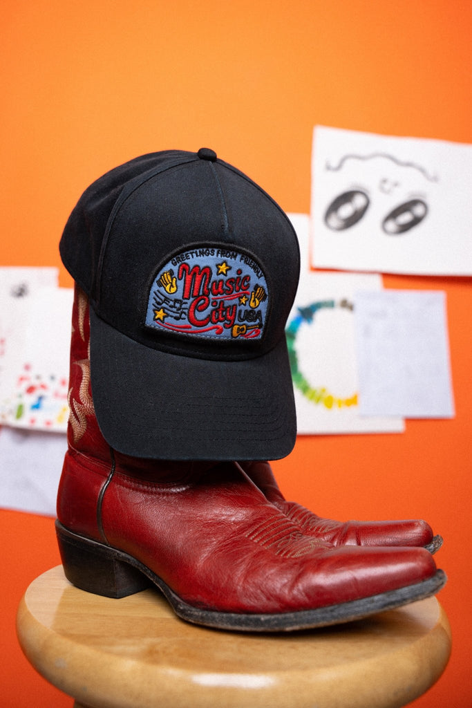 Black cap with a colorful 'Music City USA' design, on a red boot against an orange background