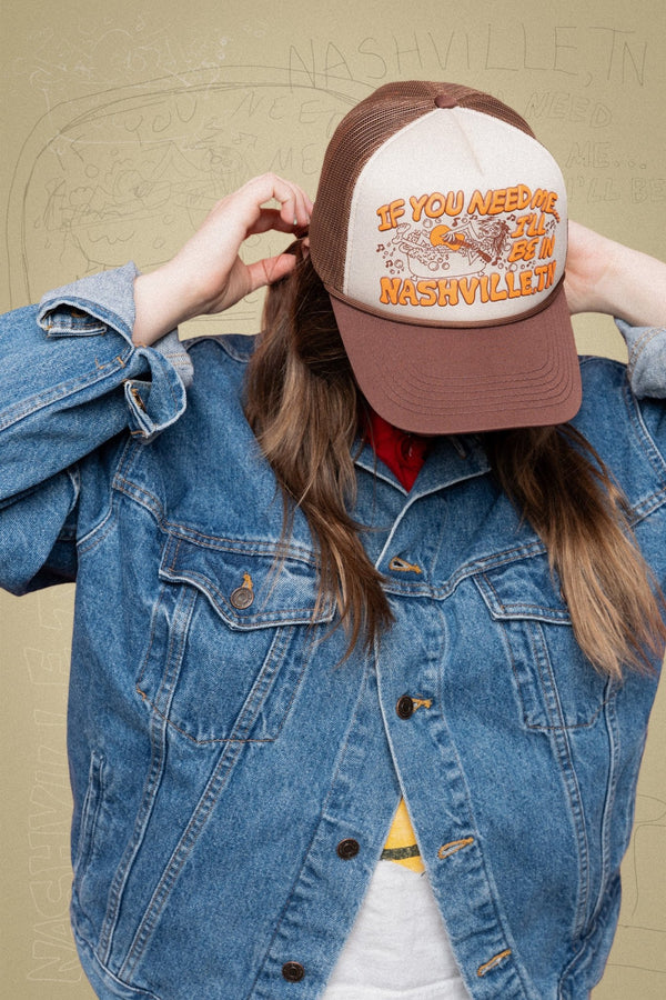 Person wearing a brown and white cap with text and graphics, that say "if you need me...I'll be in Nashville, TN" standing against a beige wall.