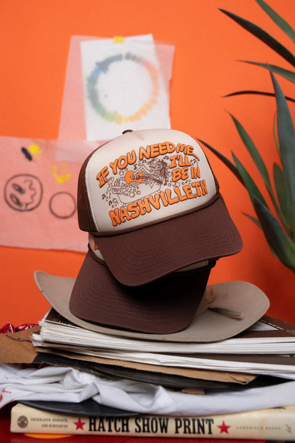 brown and white cap with text and graphics, that say "if you need me...I'll be in Nashville, TN" on a stack of books against an orange background