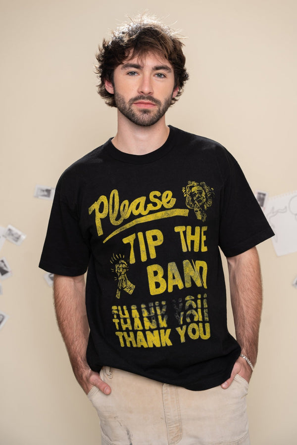 Man wearing a black t-shirt with yellow text that says "please tip the band" on a beige wall background