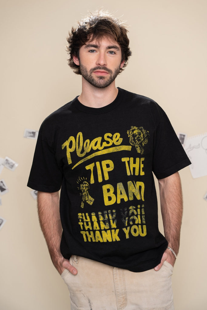 Man wearing a black t-shirt with yellow text that says "please tip the band" on a beige wall background