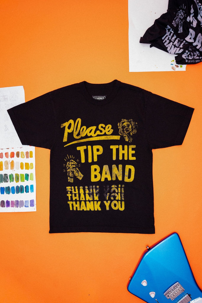 Black t-shirt with yellow text that says "Please tip the band thank you" on an orange surface