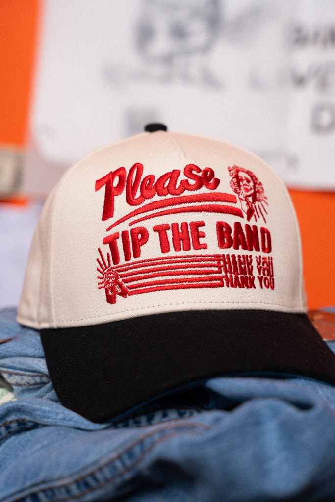 Beige and black cap with red text 'Please Tip The Band' on a denim surface.
