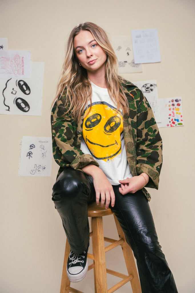 Person wearing a camouflage jacket over a white shirt with a yellow smiley face design, sitting on a stool.