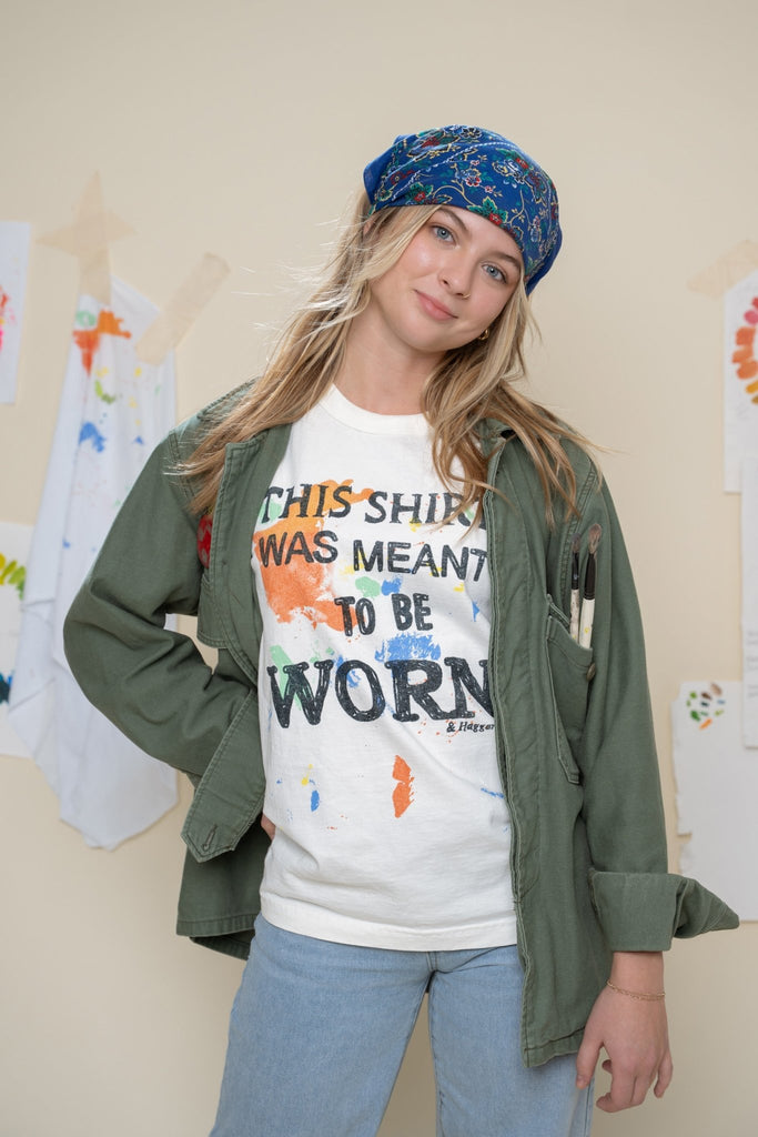 Person wearing a colorful shirt with text "this shirt was meant to be worn", and a green jacket indoors.
