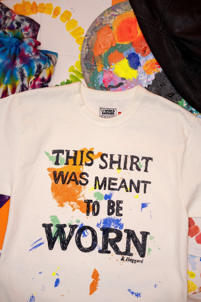 White sweatshirt with colorful text that says "this shirt was meant to be worn", and paint splashes, displayed against a colorful abstract background.